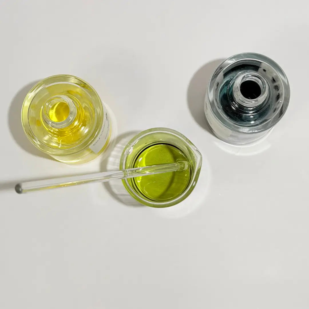 Two By Valenti face serums, Bakuchiol smoothing serum golden yellow, Olio di C face serum blue and a beaker with a green serum that combines equal parts of the other two on a white background