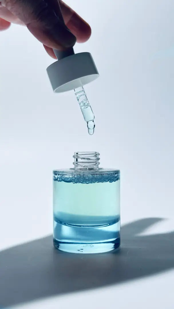 Hand holding a dropper over a bottle of By Valenti Barcola Azzurro Mare Replenishing Elixir's aqua-blue serum on a light background