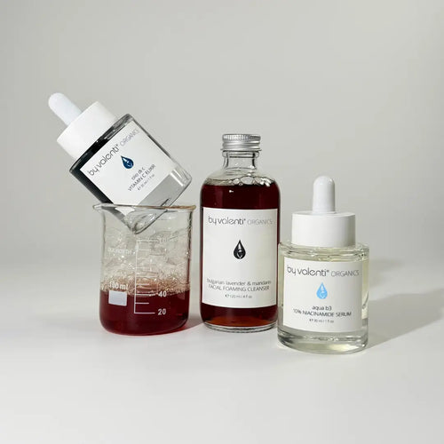 Collection of skincare products from BY VALENTI on a neutral background and a beaker containing a dark amber foaming cleanser