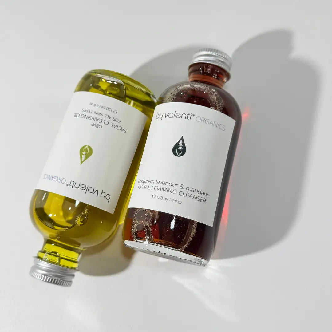 Two glass bottles of BY VALENTI lavender face soap and olive makeup remover on a white background
