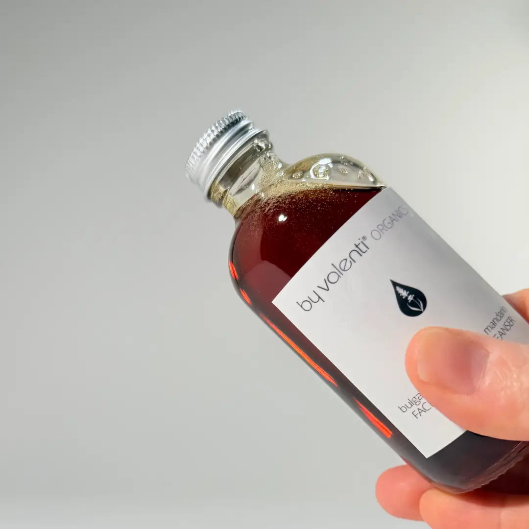 Hand holding a 120 ml glass bottle of BY VALENTI dark amber lavender and mandarin facial foaming cleanser with an aluminium cap on white background
