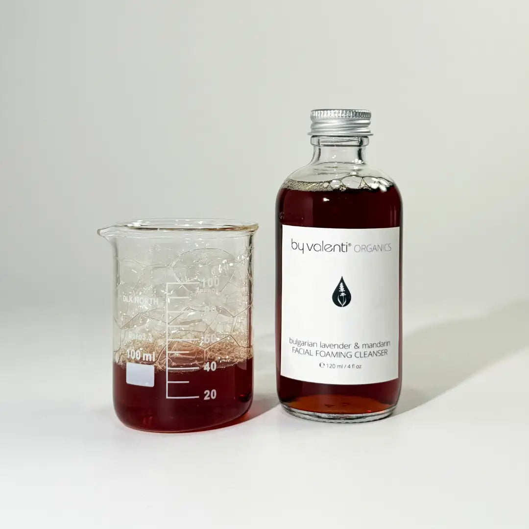 Glass bottle of BYVALENTI dark amber color lavender foaming cleanser for the face next to a beaker with some of the cleanser with bubbles on a white background