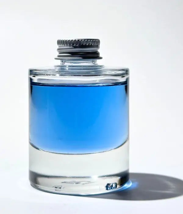 Clear glass bottle of By Valenti Olio di C Vitamin C Serum with Blue Tansy and an aluminium cap on a white background casting a shadow reflecting the blue liquid on the table