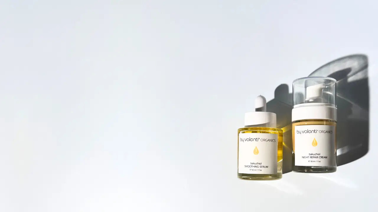 A glass bottle with dropper of by valenti golden yellow bakuchiol smoothing serum next to a glass bottle with pump of by valenti butter colored night repair cream laying flat on a light blue background casting long afternoon shadows. 