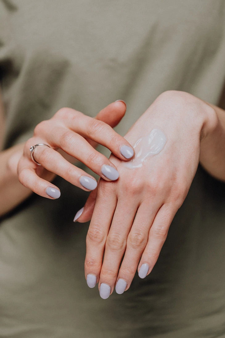 Close-up of woman's hands with light purple nail polish applying a white cream to another hand against a neutral background.