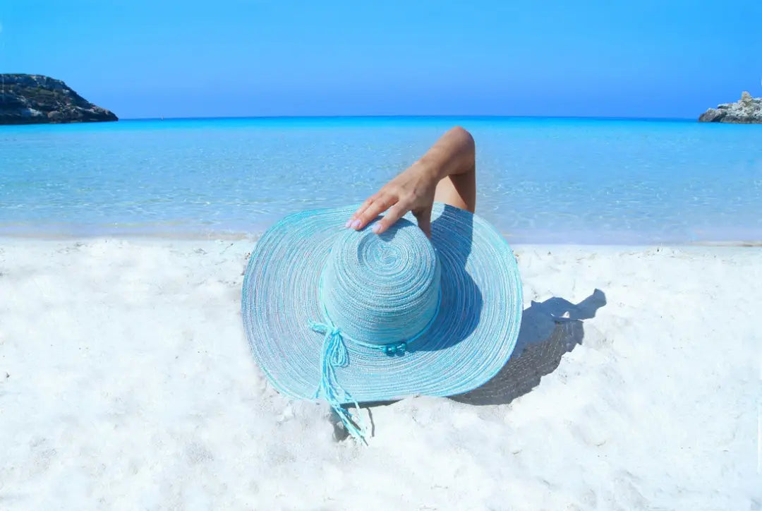 Woman wearing a large light-blue hat laying on a sandy beach with clear blue water and sky.