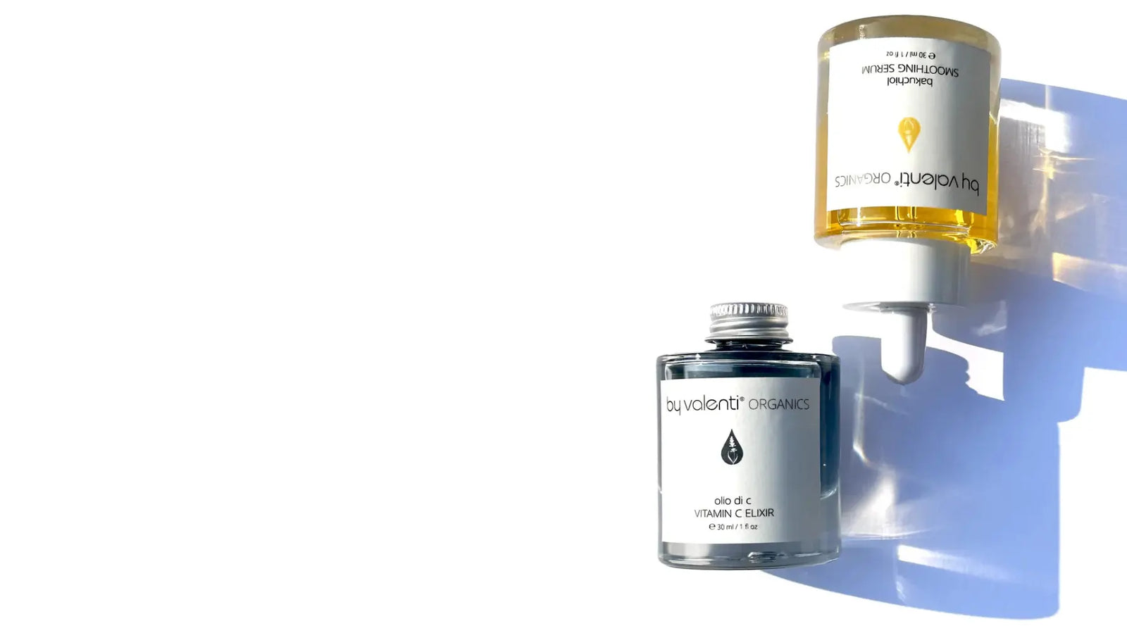 Two clear glass bottles of serums, one with a golden liquid for By Valenti Organics Bakuchiol Smoothing Serum with a white dropper cap, and the other with a blue serum with an aluminium cap, both casting a late afternoon blue-purple shadow over a white background